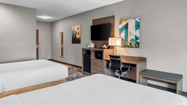 Habitación de hotel con dos camas, un escritorio con ordenador, televisor y obras de arte en la pared.