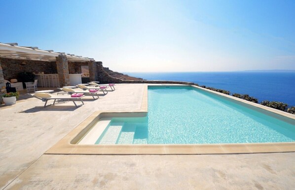 Une piscine avec vue sur la mer, des chaises longues et un bâtiment en pierre.