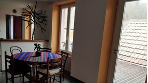 A dining area with a round table, chairs, and a colorful tablecloth. There is a potted plant on the table and a window with blinds. - Petit Appartement Lumineux et Indépendant (Lalaye)