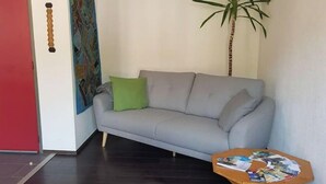 A living room with a grey sofa, a green pillow, a tall plant, and a small wooden table with magazines. - Petit Appartement Lumineux et Indépendant (Lalaye)