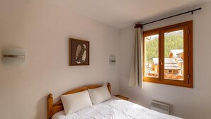 A bedroom with a wooden bed, white bedding, a framed picture on the wall, a window with curtains, and a view of a wooden cabin. - 150m from ski slopes, 4th floor, view resort, swimming pool, sauna, balcony, ski locker, 33m² (Les Orres)