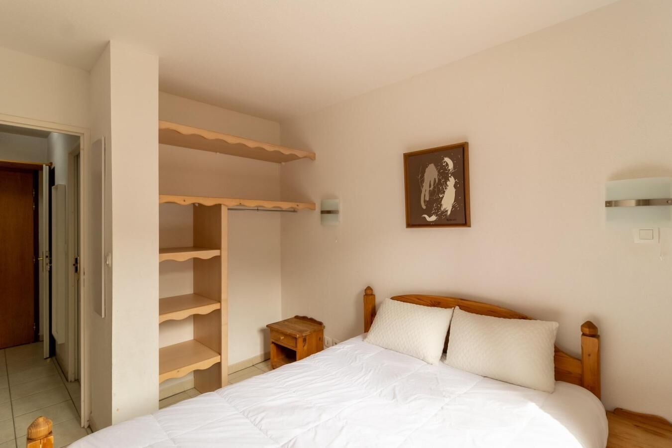 A bedroom with a bed, wooden headboard, a nightstand, a wooden wardrobe with open shelves, a framed picture on the wall, and a door leading to another room.