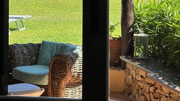 Een overdekt terras met een rieten zithoek en een tafel, met uitzicht op een grasrijk landschap en verre gebouwen aan zee.
