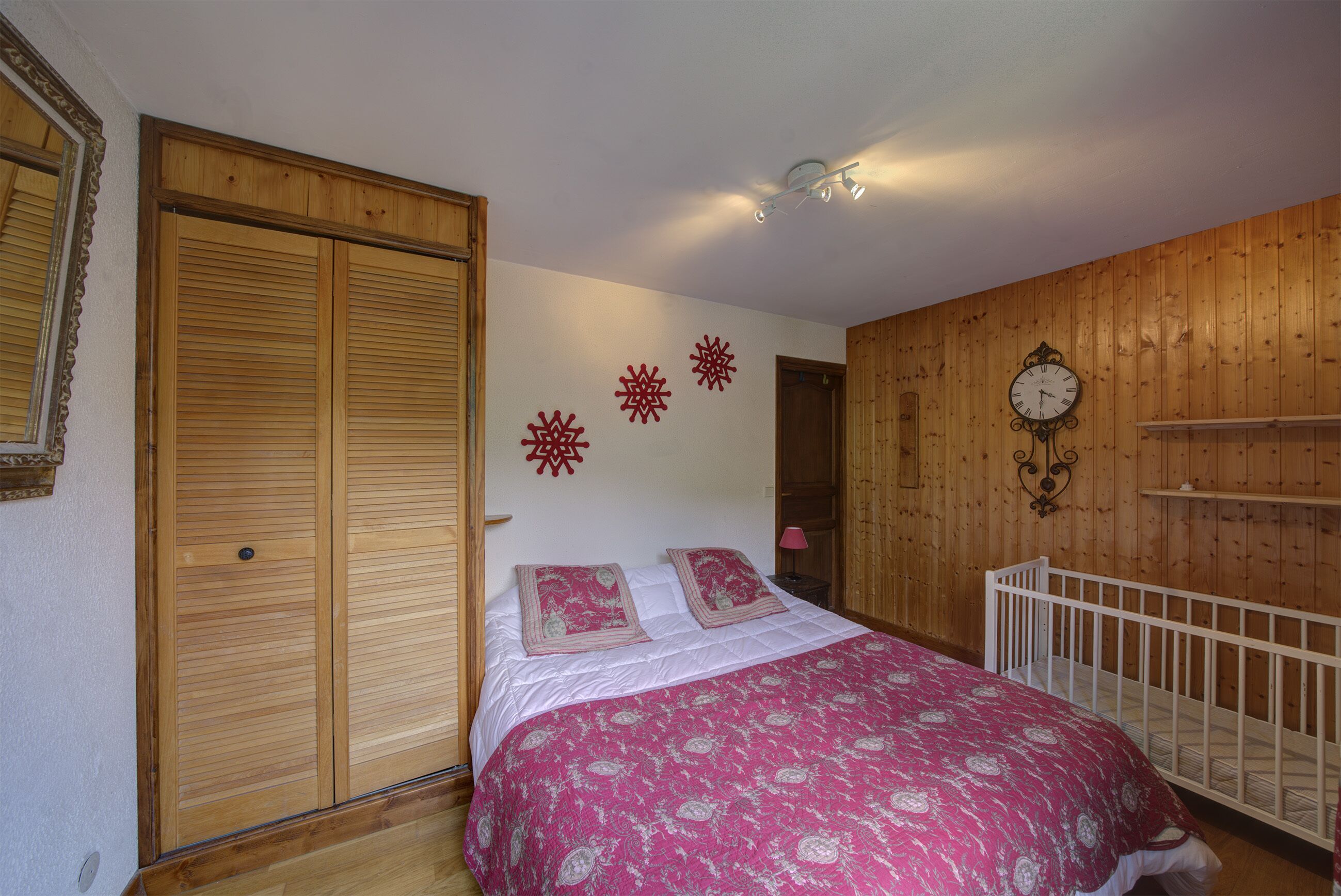 A bedroom with a wooden wardrobe, a bed with a pink patterned bedspread, a crib, and a wall clock.