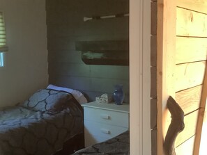 A small, dimly lit room with a single bed, a nightstand, and a wall-mounted shelf.