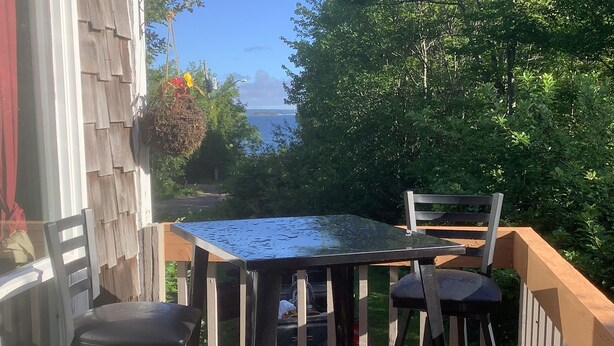 A wooden deck with a glass table and two chairs, overlooking a garden and a body of water. - Swimmin pool movie stars &a c’ment pond- jc ma gee (Shédiac)
