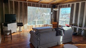 A cabin interior with a checkered sofa, wooden floors, a television, and large windows offering a view of trees and buildings. - Swimmin pool movie stars &a c’ment pond- jc ma gee (Shédiac)