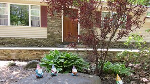 A garden with three gnome statues on a rock, a stone wall, and a house with a window and shutters. - Cozy Home With Fire Place 10 Mins From Hunter Mountain (Palenville)