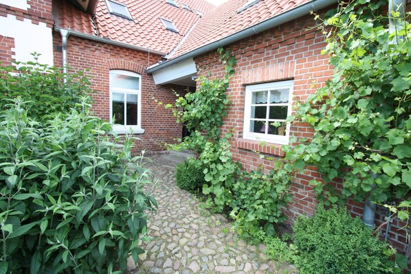 A brick house with a stone pathway leading to the entrance, surrounded by lush greenery and a red-tiled roof.