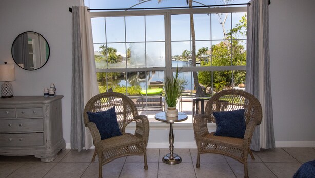 A room with two wicker chairs, a small round table, and a view of a pool and palm trees through a large window. - Ellie’s Landing 2 – Waterfront Gem w/ 2 Masters (Port Charlotte)