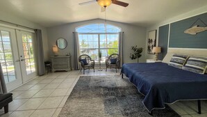 A bedroom with a large bed, a ceiling fan, a view through a window, and a seating area with a chair and table. - Ellie’s Landing 2 – Waterfront Gem w/ 2 Masters (Port Charlotte)