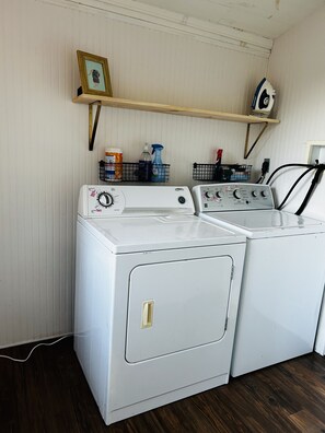 A laundry area with two front-loading washers and dryers, a shelf with cleaning supplies, and a framed picture on the wall. - Adorable private cottage with lake views and access. (Dedham)