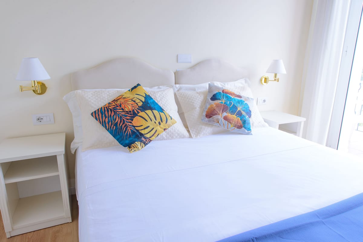 Uma cama com roupa de cama branca e duas almofadas decorativas, uma mesa de cabeceira e uma luminária fixada na parede.