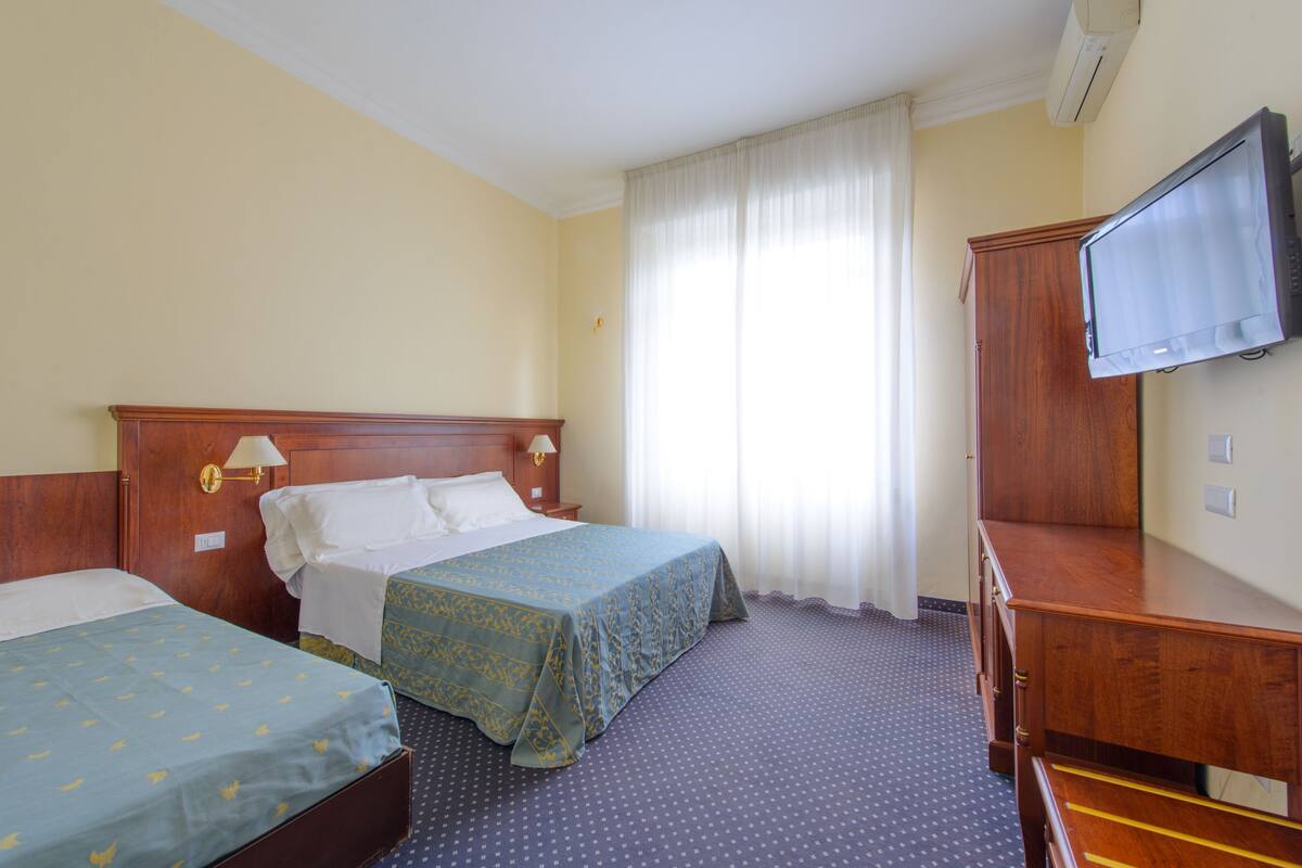 Quarto de hotel com duas camas, um guarda-roupa de madeira, uma televisão e uma janela com cortinas.
