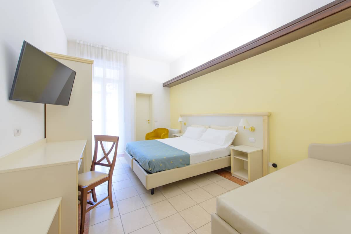 Quarto de hotel com cama, escrivaninha, cadeira, televisão e mesa de cabeceira.
