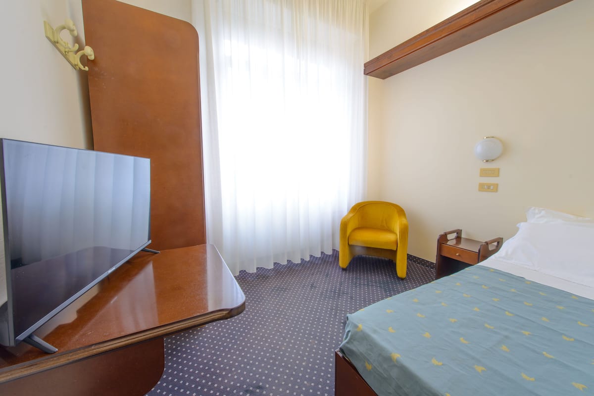 Quarto de hotel com televisão de tela plana, cama com colcha azul, poltrona amarela, mesa de cabeceira de madeira e luminária na parede.