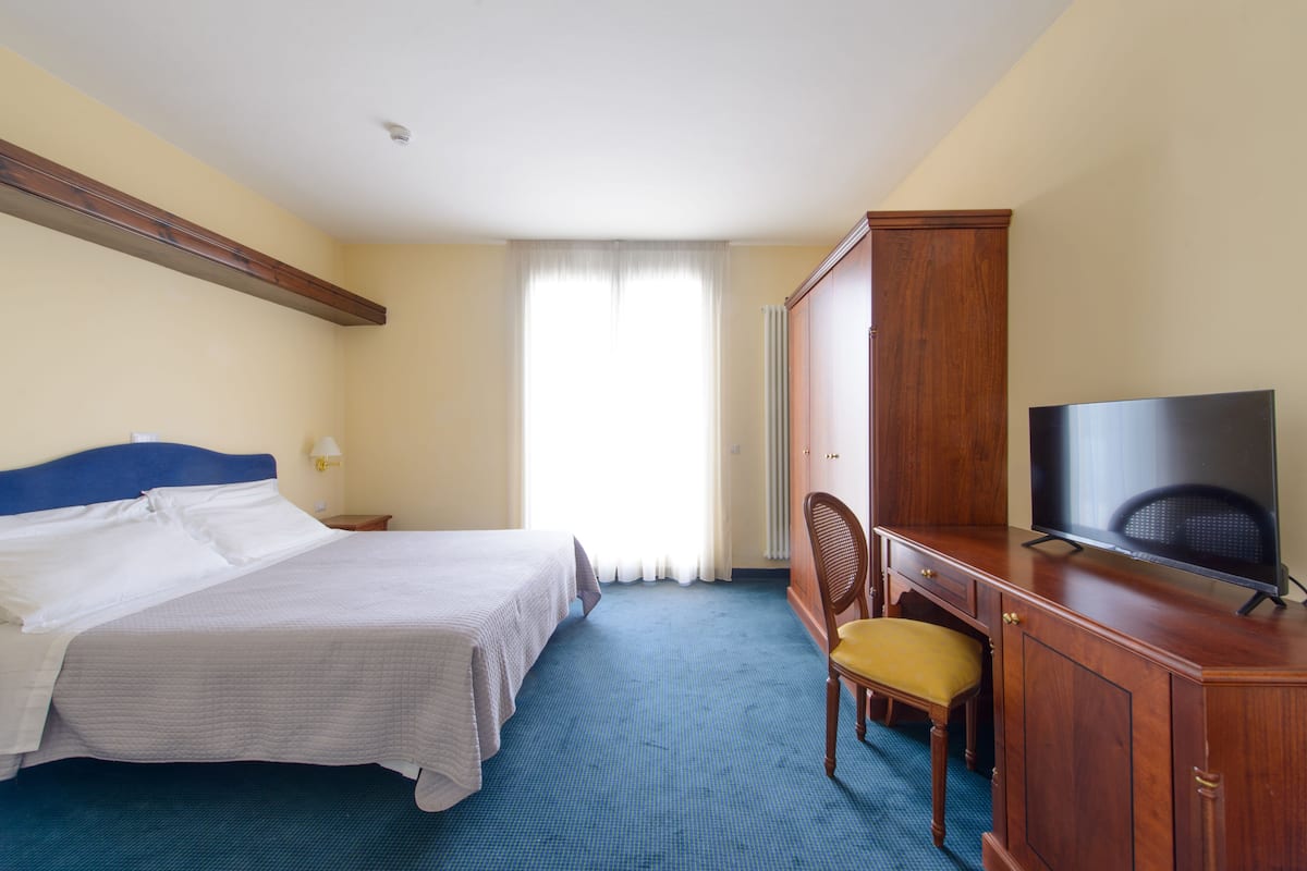 Quarto de hotel com cama, televisão, guarda-roupa de madeira, uma cadeira e janela com cortinas.