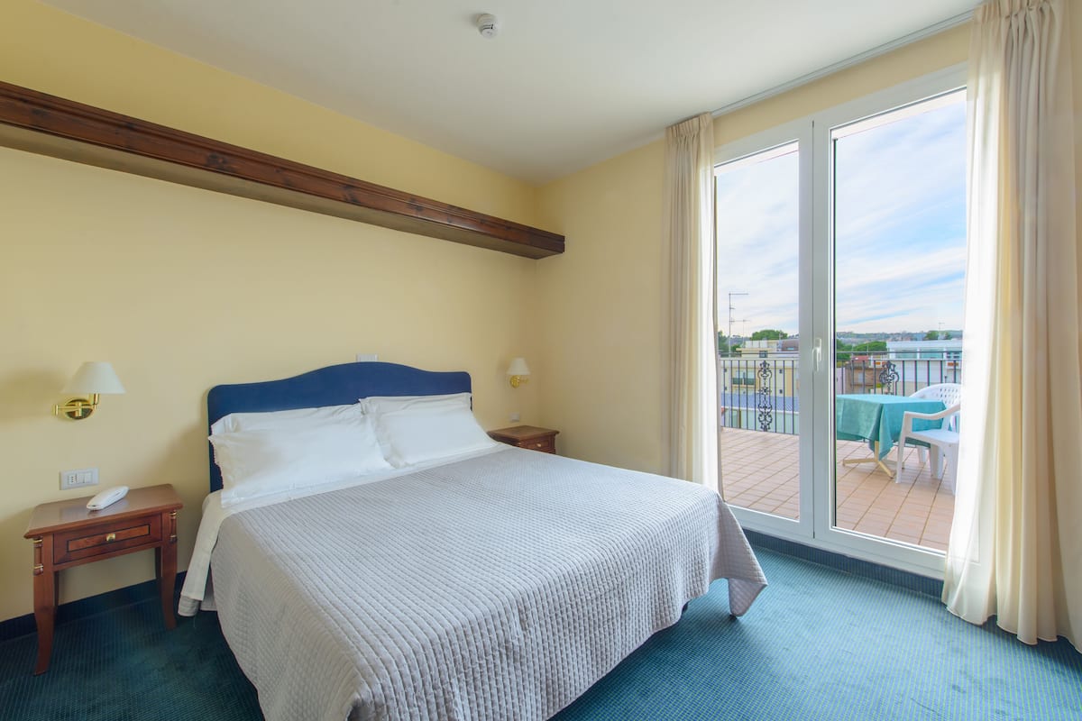 Quarto de hotel com uma cama grande, duas mesas de cabeceira, uma janela com vista para o exterior e uma varanda com mesa e cadeiras.