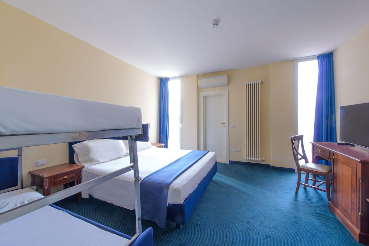 Quarto de hotel com beliches, uma escrivaninha, uma cadeira, uma televisão e uma janela com cortinas.