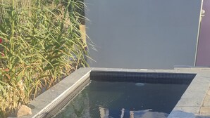 Uma piscina com borda azulejada, rodeada por pedras e plantas.