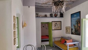 Um quarto com uma mesa de jantar, uma cama com colcha estampada em amarelo e vermelho, uma porta verde e um pôster de 'La Cenerna di Tito' na parede.