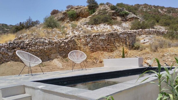 Uma área moderna de piscina ao ar livre, com duas cadeiras brancas, um muro de pedra e uma escadaria que leva a um nível superior.