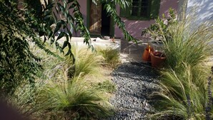 Um caminho no jardim leva a um edifício com persianas verdes e uma parede rosa.