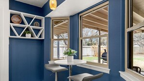 A corner room with a blue wall, a round table with two bar stools, a window with a view of the backyard, and a shelf with decorative items.