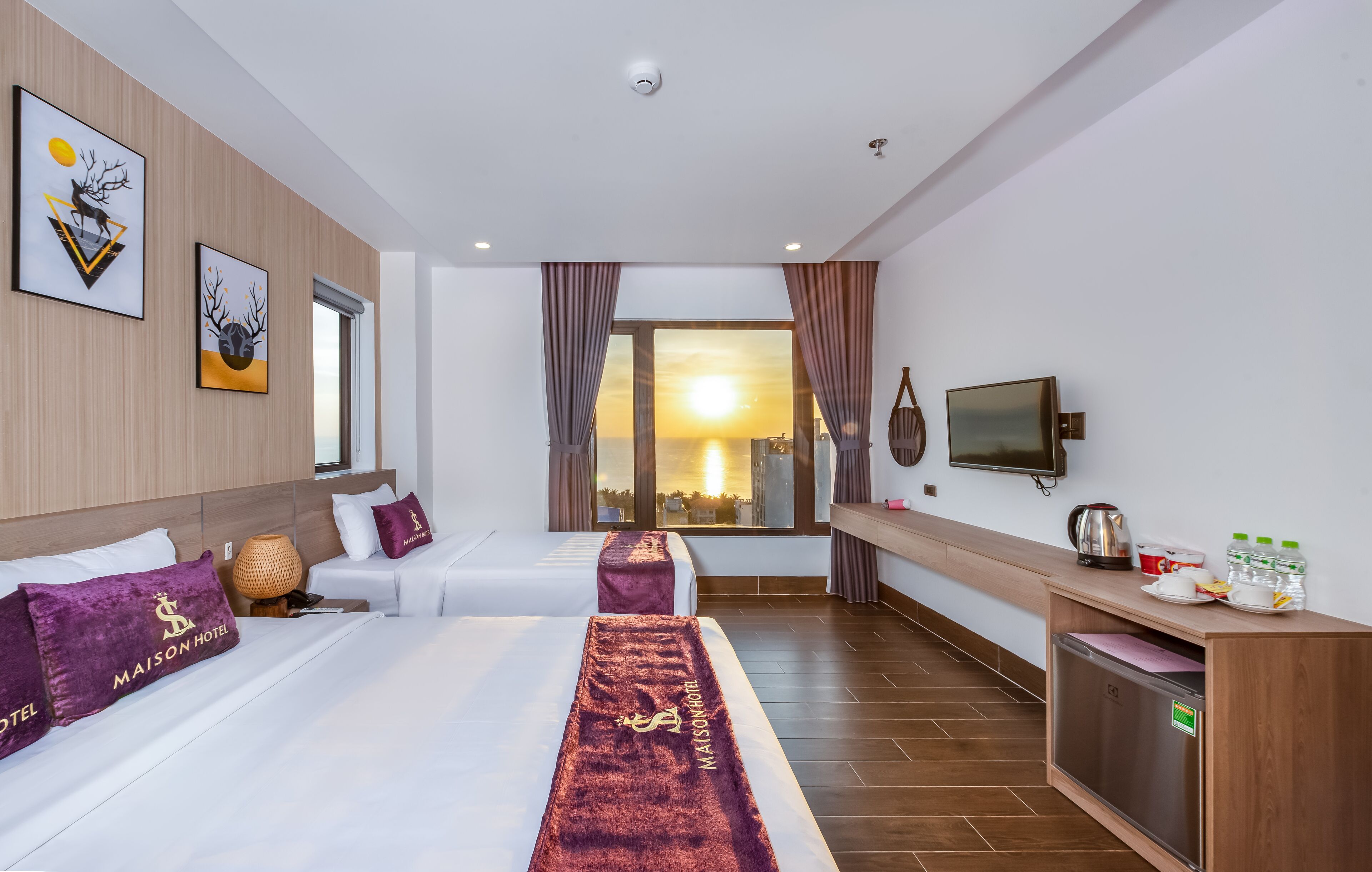 A hotel room with two beds, a TV, a minibar, and a large window with a sunset view.