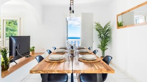 A dining area with a wooden table set for six, black chairs, a TV on the left, and a view of the sea through a window. - Family villa with private heated pool and ocean views (Tabaiba)