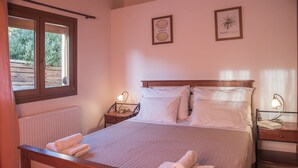 Una cama bien hecha con sábanas blancas y dos toallas enrolladas, una mesita de noche con una lámpara e impresión enmarcada de motivos botánicos en la pared.