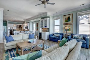 A spacious living room with a large sofa, a coffee table, and a blue armchair. There is a kitchen area with white cabinets and a dining table. - Tipsy Turtle North- Oceanfront Oasis - Pool - Dog Friendly (Surf City)