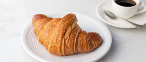 Een croissant op een wit bord, met een kop koffie en een lepel op een schoteltje.