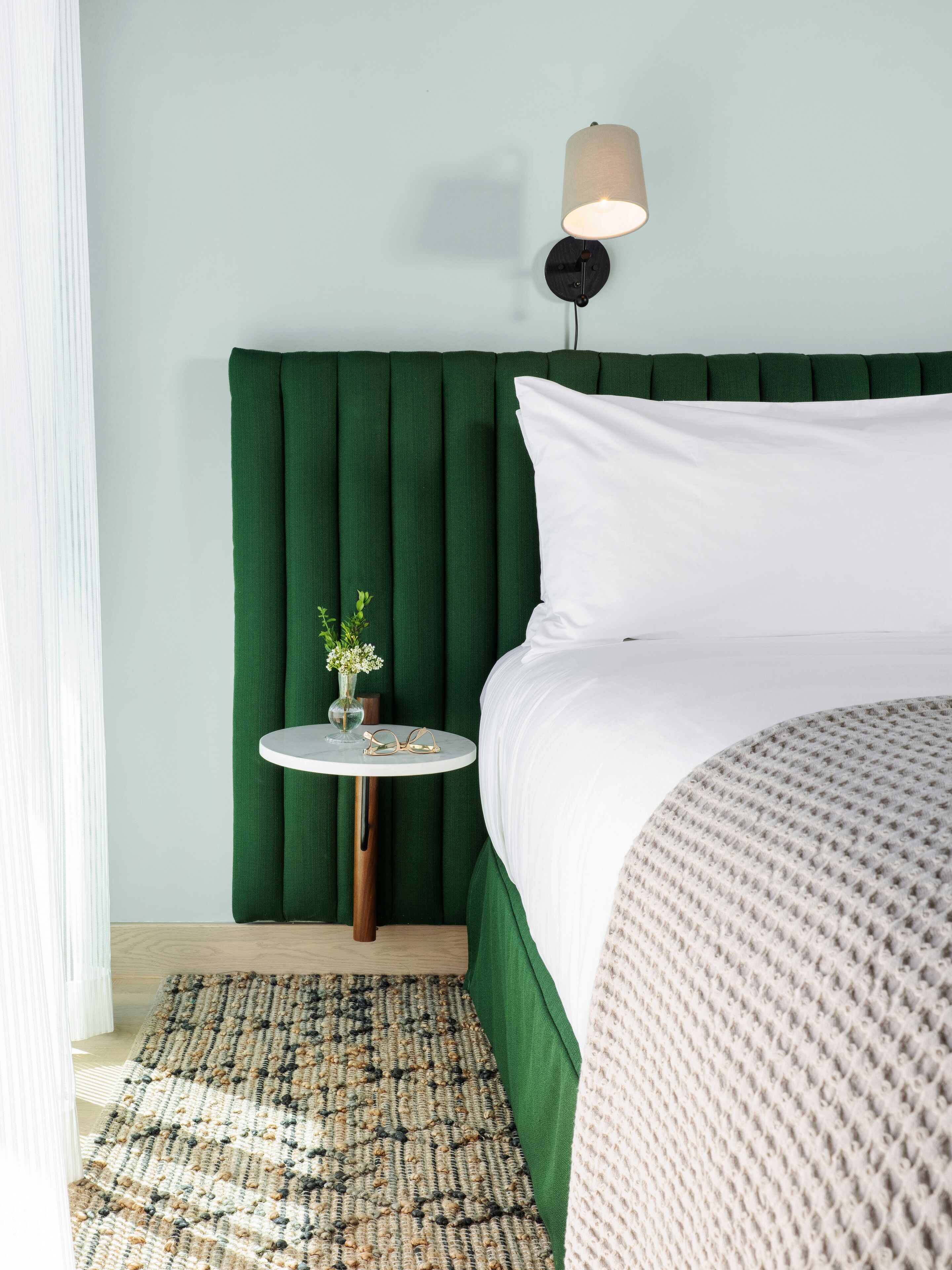 A neatly arranged hotel room with a green upholstered headboard, a round bedside table with a vase and glasses, and a bed with a white pillow and a textured blanket.