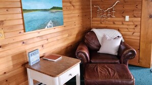 Une pièce chaleureuse aux murs en bois, avec un fauteuil en cuir marron, une petite table blanche et un tableau représentant un paysage.