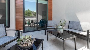 Ein moderner Außenbereich mit Glastischen und Metallstühlen, einer Pflanztopf und Blick auf eine Terrasse mit Gartenmöbeln.
