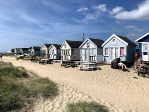 Een rij strandhuisjes met buitenmeubilair en een zandstrand op de achtergrond.