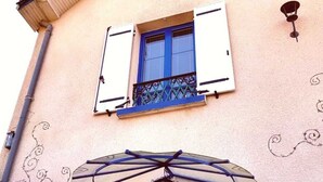 Un edificio con techo a dos aguas, una ventana con contraventanas azules, un balcón decorativo y un farol de pared.