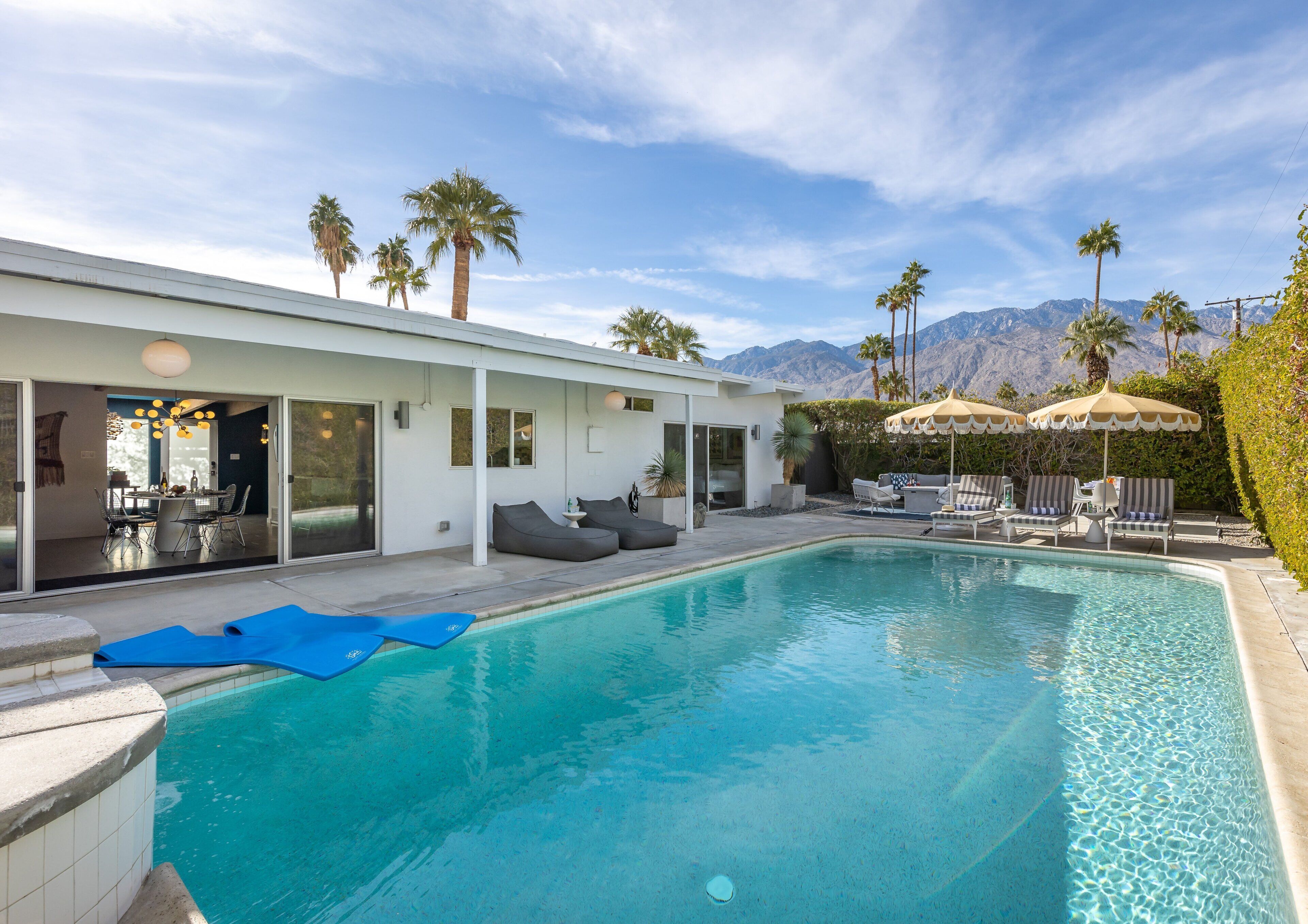 A modern pool area with a clear view of mountains, a dining area with a table and chairs, and a lounge with sun loungers.