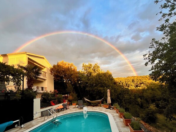 Piscina con sillas de descanso y una hamaca, rodeada de árboles y un edificio.