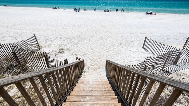 Ein hölzerner Steg führt zu einem Sandstrand, im Hintergrund liegt klares, blaues Wasser.