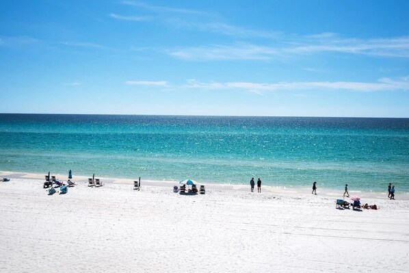 Ein Strand mit klarem, blauem Wasser, weißem Sand und mehreren Liegestühlen und Sonnenschirmen.