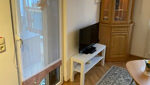 A room with a sliding glass door, a television on a white stand, a small white table, a patterned rug, and a wooden cabinet with glass doors. - Ferienwohnung Greiz W2 (Greiz)
