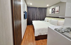 A modern hotel room with a large bed, a bedside table with cups, a flat-screen TV, and a large window with curtains.
