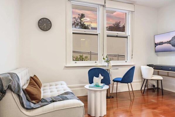 A modern living room with a sofa, two chairs, a small table, and a large window with a view of trees and a sunset.
