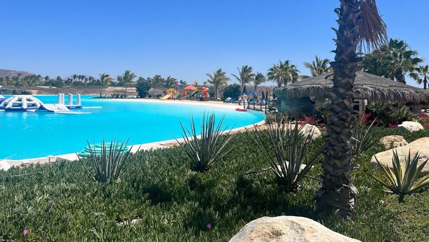 Una piscina azul cristalina con un tobogán y sombrillas de techo de paja, rodeada de palmeras y rocas.