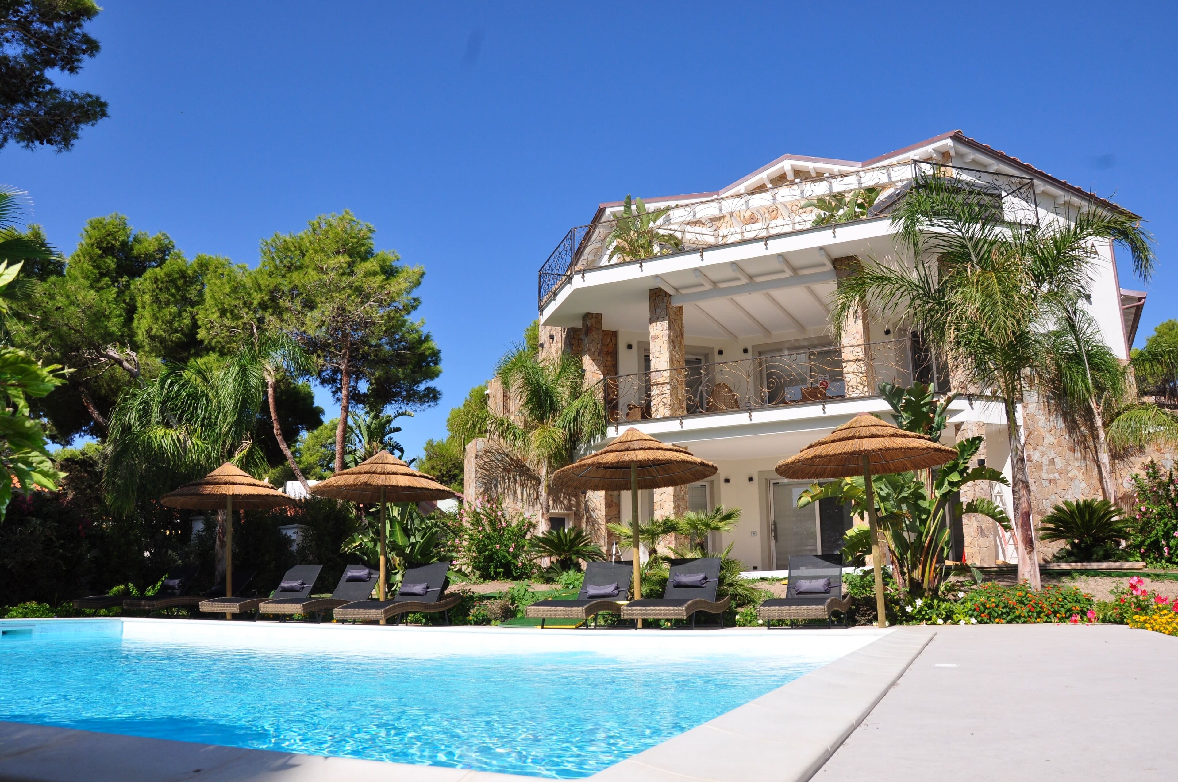A poolside area with lounge chairs and umbrellas, a clear blue pool, and a large building with columns and a balcony.