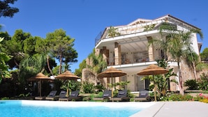 A poolside area with lounge chairs and umbrellas, a clear blue pool, and a large building with columns and a balcony.