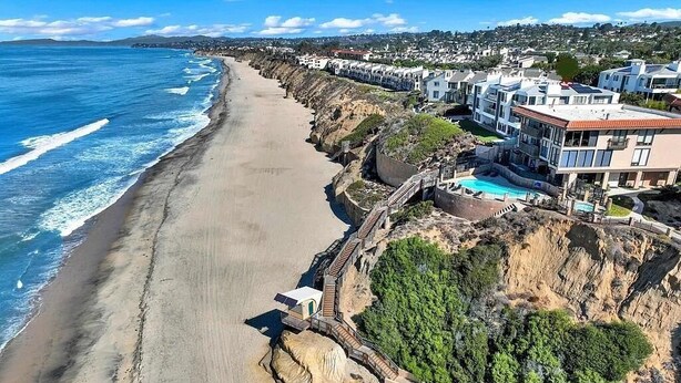 Vista aerea di una zona costiera con spiaggia sabbiosa, una passerella in legno e edifici residenziali con balconi e una piscina.
