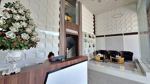 Zona d'entrada d'un hotel amb disseny modern, que destaca un taulell de recepció de fusta, una planta decorativa i una escala que condueix a un pis superior.